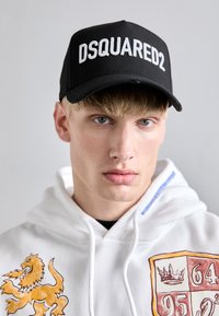 Gorra negra con el texto en blanco "DSQUARED2" en la parte frontal, llevada con una sudadera blanca que presenta un gráfico de león y un diseño de emblema.