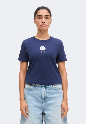 Žena s tmavými vlasmi stiahnutými dozadu, nosi tmavomodré tričko s krátkym rukávom s logom Paríž a bielym logom Nike, v kombinácii so svetlomodrými džínsami.