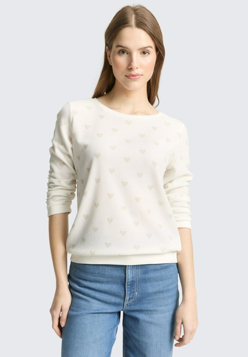 Giovane donna con camicia bianca a maniche lunghe con sottili motivi a cuore e jeans blu, in piedi contro uno sfondo chiaro e semplice.