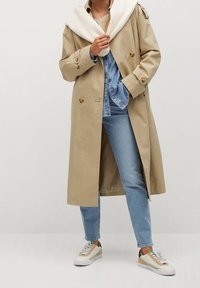 Trench-coat beige à double boutonnage, associé à un jean en denim clair et des baskets multicolores, avec des lacets blancs et des accents verts.