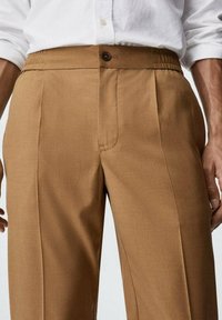 Braune, plissierte Hose mit gewobener Textur, mit einem Knopfverschluss an der Taille und seitlichen Taschen. Das Material wirkt langlebig und strukturiert.