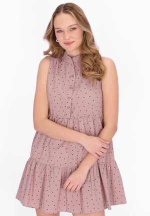 Junge Frau mit langen, welligen Haaren, die ein ärmelloses, rosa Kleid mit schwarzen Punkten trägt, die Arme locker vor der Taille verschränkt.