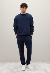 Sudadera de color azul marino con un logo "P" texturizado en el pecho, combinada con joggers a juego que tienen bolsillos frontales, complementada con zapatillas blancas.