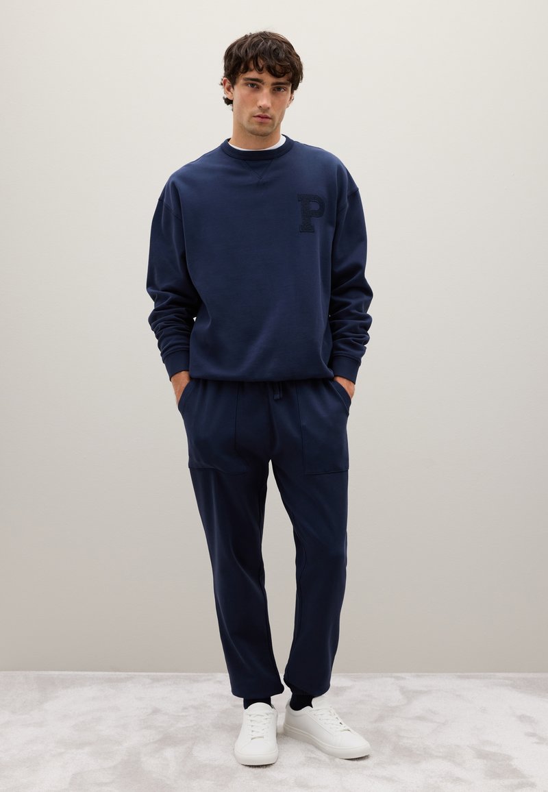 Sudadera de color azul marino con un logo "P" texturizado en el pecho, combinada con joggers a juego que tienen bolsillos frontales, complementada con zapatillas blancas.