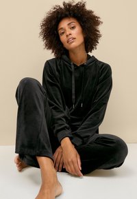Zwarte velours loungeoutfit bestaande uit een hoody en bijpassende broek. Soepele textuur en ontspannen pasvorm, met trekkoorden in de capuchon.