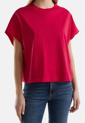 Kobieta ubrana w luźną, jasnoczerwoną bluzkę z krótkim rękawem oraz niebieskie dżinsy, stojąca na tle jasnego, jednolitego tła.