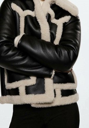Personne portant une veste en cuir noire avec une bordure en shearling couleur crème, bras croisés, sur un fond clair uni.