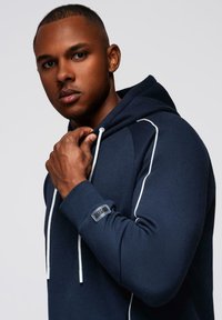 Zbliżenie na mężczyznę w granatowym kapturze z białymi sznurkami, trzymającego kołnierz blisko podbródka, patrzącego prosto przed siebie.