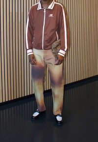 Hnědá bunda New Balance na zip s bílými pruhy; světle hnědé kalhoty s přechodem barev; černo-bílé boty; doplněk v podobě kožešinové kapsičky.