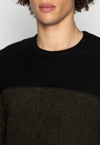 Stickad tröja med texturerad svart överdel och olivgrön underdel, med rund halsringning och ribbad krage.