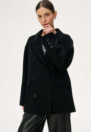 Kobieta z zaczesanymi do tyłu włosami nosi czarny dwurzędowy płaszcz na wzorzystej bluzce i czarnych skórzanych spodniach, pozując z ręką przy twarzy.