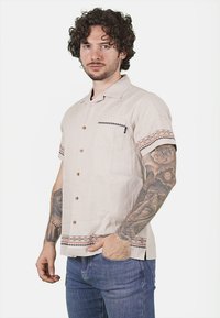 Camicetta beige a maniche corte realizzata in tessuto leggero, con taschino sul petto e dettagli decorativi sulle maniche e sull'orlo.