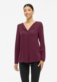 Blusa bordeaux realizzata in tessuto liscio, con scollatura a V, maniche lunghe e orlo dritto con sottili spacchi laterali.