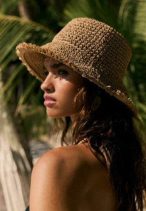 Kobieta o długich ciemnych włosach, nosząca szeroki słomkowy kapelusz z plecionki, patrząca w bok, na tle rozmytych zielonych liści palm.