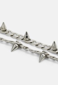 Accesorio de cadena plateada que presenta dos filas: una con picos y otra torcida, ambas exhibiendo un acabado metálico brillante.