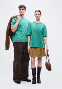 Νεαρός άνδρας και γυναίκα στέκονται δίπλα-δίπλα, και οι δύο φορούν μπλουζάκια Tommy Jeans σε τιρκουάζ χρώμα· ο άνδρας κρατά καφέ μπουφάν, η γυναίκα καφέ τσάντα χειρός.