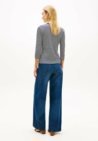 Kobieta o długich blond włosach, ubrana w granatową bluzkę w paski z rękawami do łokci i szerokie niebieskie dżinsy, stoi tyłem na białym tle.