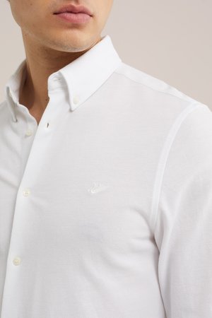Uomo che indossa una camicia bianca con bottoni, colletto abbottonato e un logo ricamato sobrio sul petto, mostrato dal collo fino a metà torace.