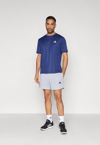 Ανδρικός αθλητικός εξοπλισμός με μπλε σκούρο T-shirt που απομακρύνει την υγρασία και ανοιχτό γκρι σορτς με μαύρες λεπτομέρειες και λογότυπο Adidas, σε συνδυασμό με μαύρα αθλητικά παπούτσια.