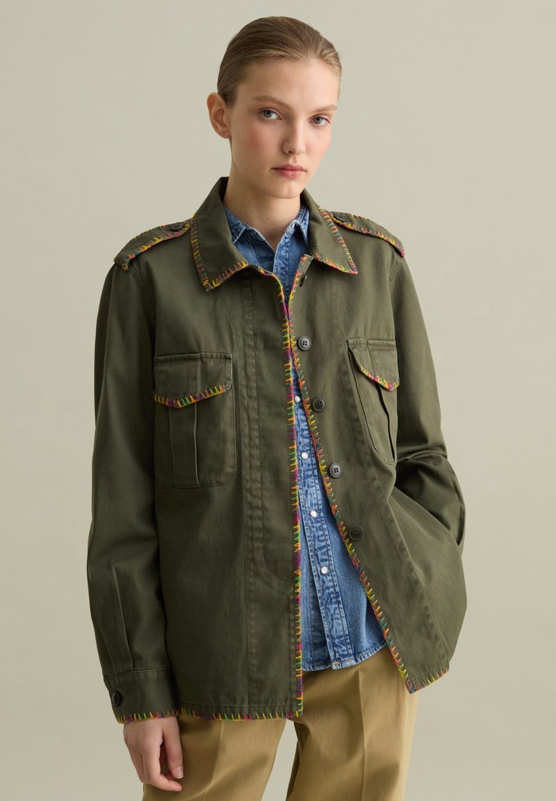 Giacca militare verde oliva con cuciture multicolori lungo il collo e le tasche, dotata di chiusura con bottoni e tasche sul petto.