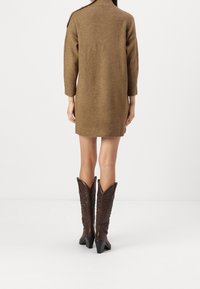 Abito maglione marrone con collo alto e maniche a tre quarti, con dettagli di bottoni sulle spalle. Abbinato a stivali cowboy marroni.