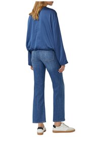 Kobieta w luźnej, niebieskiej bluzie z długim rękawem, prosto skrojonych niebieskich dżinsach i białych trampkach z czarnymi akcentami na pięcie, stojąca tyłem.