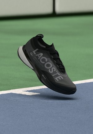 Sort Lacoste sportsko med stort logo, svævende over en blå og grøn tennisbane.
