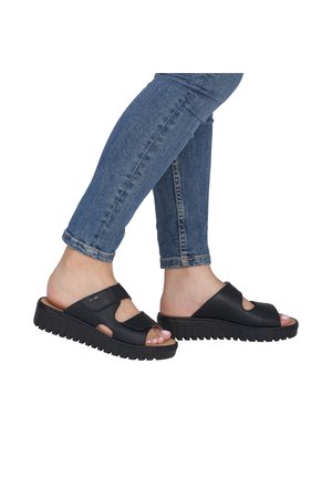 Feet wearing black slip-on sandals with thick soles paired with blue rolled-up skinny jeans against a white background.