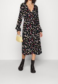 Černé šaty s kapsou evokující listy v barevných vzorech, dlouhé rukávy s volánky, nošeno s černými kotníkovými botami a malou žlutou kabelkou.