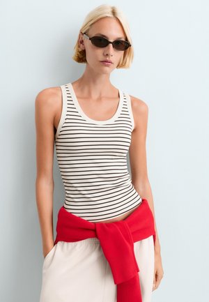 Femme aux cheveux blonds courts portant des lunettes de soleil noires étroites, un débardeur blanc à fines rayures noires et un pull rouge noué autour de la taille.