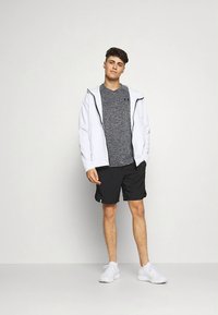 Bela jakna z zadrgo čez sivo kratko majico, črni kratki hlači in beli supergi. Preprost dizajn brez vidnih vzorcev ali poudarkov.