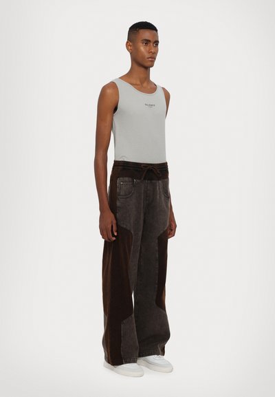 Grijze tanktop met 'SILENCE' logo, gecombineerd met losse, donkergrijze jeans met bruine corduroy accenten en een elastische tailleband. Witte sneakers.