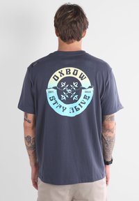 T-shirt en coton bleu marine avec un grand graphique circulaire comportant le texte "OXBOW" et un design de montagne en jaune et bleu clair. Manches courtes.