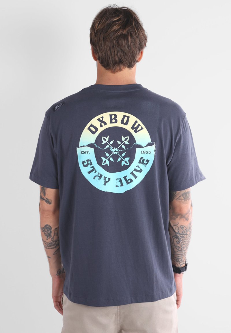 T-shirt en coton bleu marine avec un grand graphique circulaire comportant le texte "OXBOW" et un design de montagne en jaune et bleu clair. Manches courtes.