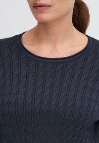 Maglione blu navy lavorato a maglia con un motivo a trecce testurizzate, scollatura rotonda e bordi a costine. Il materiale sembra morbido e leggermente elasticizzato.
