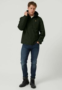 Chaqueta impermeable de color verde oscuro con un alto cuello, cierre de cremallera y logo bordado. Llevada sobre vaqueros azules y botas negras. Textura suave.