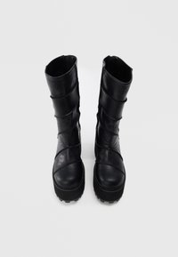 Un par de botas negras de media caña con paneles texturizados, punteras redondas y suelas gruesas, de pie sobre un fondo blanco liso.