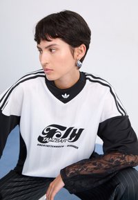 Melnas un baltas svītrots sporta krekls ar mežģīņu piedurknēm, priekšā ir grafisks uzraksts un logotips. Modelim ir īsie mati un auskari.
