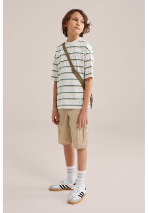 Berniukas, vilkintis dryžuotą per didelį marškinėlį, smėlio spalvos krovinių šortus, baltus sportbačius su juodomis juostelėmis, baltas kojines ir žalią per petį nešiojamą krepšį, stovintis priešais paprastą foną.