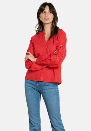 Tume juustega naine, kes kannab kuldseid kõrvarõngaid, punast pluusi tikandiga varrukalõigetega ja siniseid teksaseid, seisab käed rüpes.