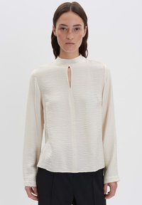 Donna con capelli scuri e lisci, che indossa una blusa lunga a maniche con scollo a chiave davanti di colore crema e pantaloni neri, in piedi contro uno sfondo semplice.