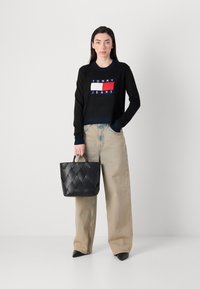 Une femme se tient debout avec un pull court noir affichant "TOMMY JEANS" à l'avant, tenant un sac à main tressé noir, portant un jean large clair.