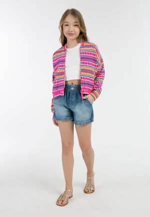 Farverig bomberjakke med mønster i pink, orange og blå over en hvid cropped top, parret med denimshorts og guld sandaler.