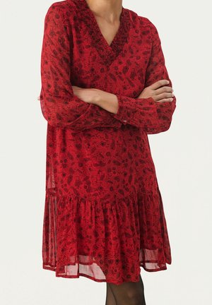 Femme portant une robe longue en fleurs rouge avec un col en V et un ourlet à volants, bras croisés sur la poitrine.