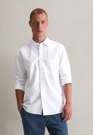 Weißes Hemd mit Knöpfen aus Baumwolle und einer Brusttasche. Lange, hochgekrempelte Ärmel, lässige Passform und subtiler Textur. Getragen mit blauen Hosen.
