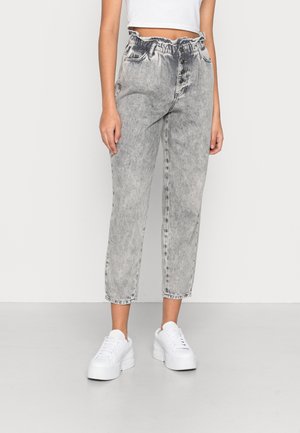 Vrouw draagt hoge taille grijze acid-wash jeans met gerimpelde tailleband en knoopsluiting, gecombineerd met witte platform sneakers.