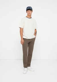White short-sleeve t-shirt with a blue ribbed neckline, brown trousers, and white sneakers. Model wears a blue cap and stands on a plain background.