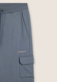Pantalons de jogging bleu clair avec taille élastique, poches latérales et poche cargo. Présente un petit logo argenté "FREDDY". Texture de tissu douce.