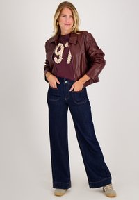 Bordeaux lederen jas, kastanjebruine t-shirt met bloemenprint, klassieke blauwe wijde jeans, grijze sneakers, zichtbare zakken en stikdetails.