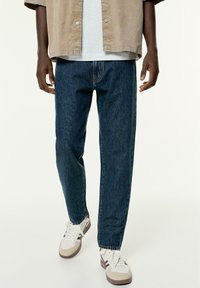 Jean en denim bleu foncé avec une coupe droite, associ�é à un t-shirt de couleur claire et des baskets beiges, blanches et marron.